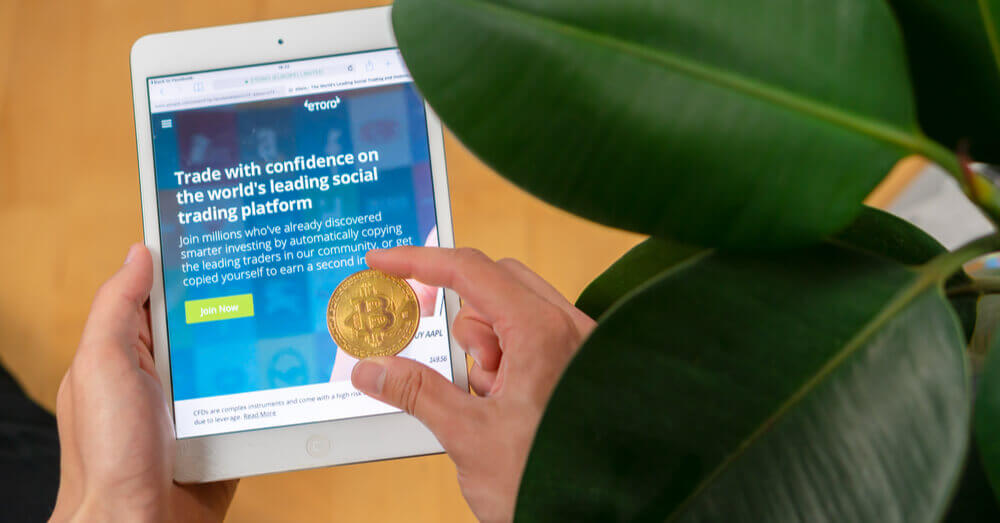 Le site d'eToro sur une tablette et une main tenant un jeton Bitcoin